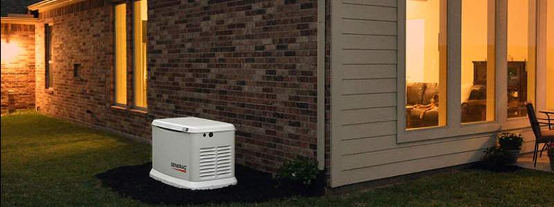 A Generac Guardian Generator next to the brick wall of a home. Inside lights cast a warm, cozy glow. To the right is a patio with table and chairs.