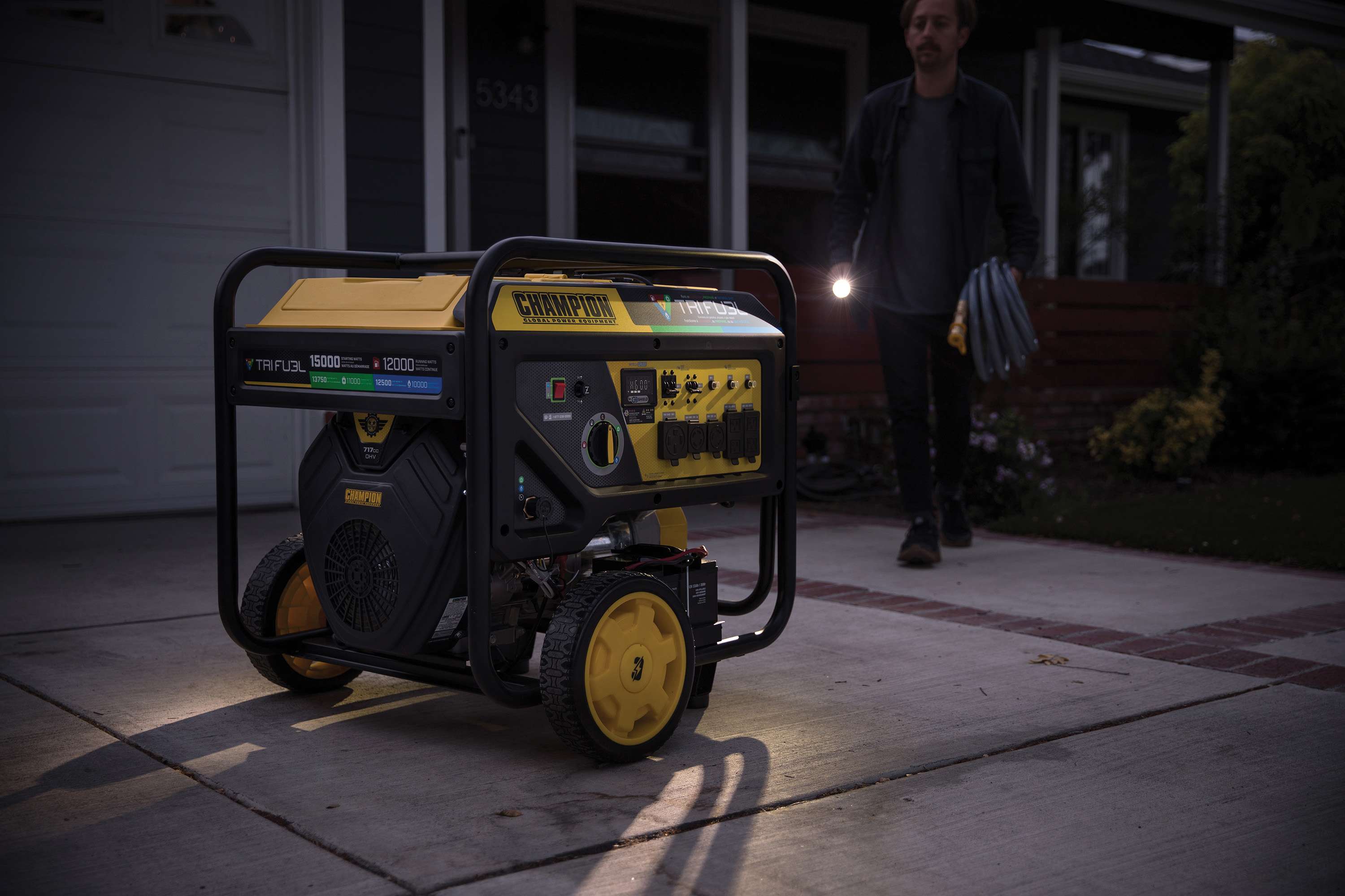 Champion 12000 Watt Generator on a driveway at night. A man approaches from a dark house carrying a flashlight and generator cord to connect the tri-fuel to the house inlet box.