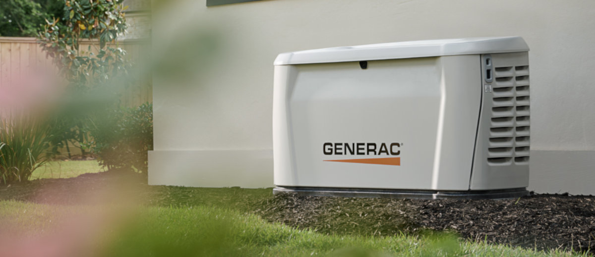 A Generac Next Gen standby generator installed near a home on a mulch bed surrounded with grass in front and shrubs in the background.