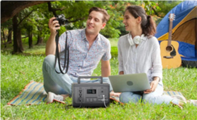 A couple sits on a blanket. He is holding a camera, she has a laptop. They are looking at pictures on the camera. There's a tent with a guitar leaning against it and trees in the background.